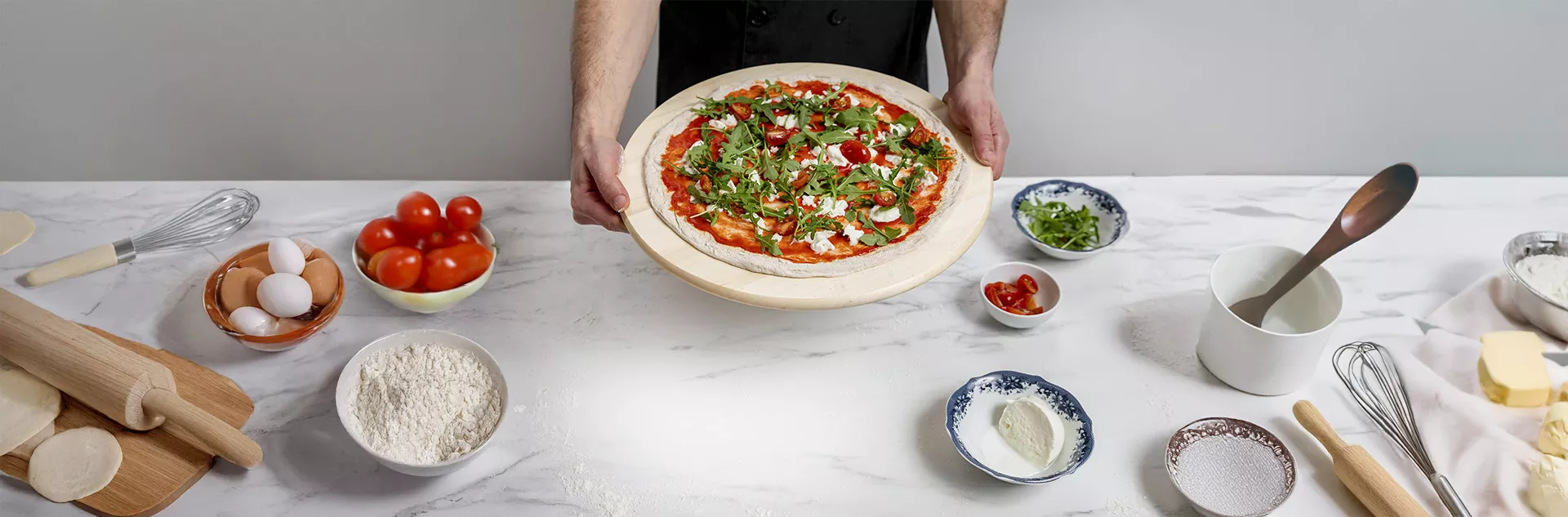 Pizzaiolo segurando pizza artesanal pronta, com rúcula, tomate-cereja e muçarela de búfala sobre bancada com ingredientes e utensílios — simbolizando padronização de receitas e controle de insumos com o CNM para pizzarias